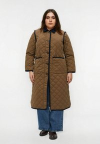 Manteau long matelassé marron avec des bordures bleu marine, fermeture à bouton et deux poches latérales. Stylé avec un pantalon en denim large et des chaussures brunes.