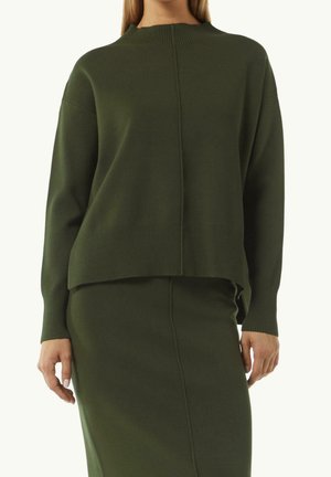 Femme portant un pull en maille côtelée vert olive à manches longues et une jupe assortie longueur genou, debout devant un fond uni.