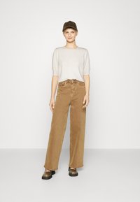 Beige svetr na krátký rukáv, hnědé široké manšestrové kalhoty, černé platformové boty a tmavá čepice; minimalistický design s jemnými texturami.