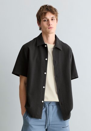 Junger Mann mit kurzen braunen Haaren trägt ein schwarzes, kurzärmliges Hemd über einem weißen T-Shirt und hellblaue, mit Kordelzug versehene Hosen vor grauem Hintergrund.