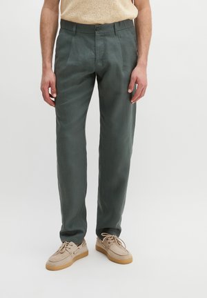 Man draagt donkergroene rechte broek, beige gebreide top en beige veterschoenen, staande op een witte achtergrond.