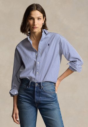 Kvinne med skulderlangt mørkt hår iført en blå og hvit stripete knappeskjorte og høytlivs blå jeans, poserer med én hånd på hoften.