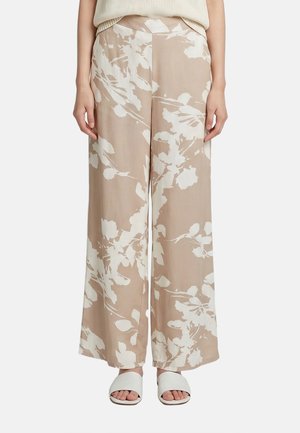 Pantaloni beige a gamba larga con stampa floreale bianca, indossati con sandali bianchi aperti davanti, su sfondo semplice.