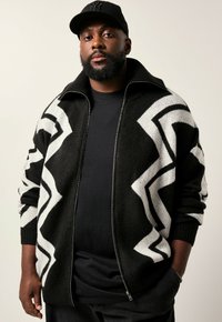 Schwarz-weißer Reißverschluss-Pullover mit eckigem Muster, gestrickter Textur, hohem Kragen und gerippten Bündchen; über einem schwarzen T-Shirt getragen.