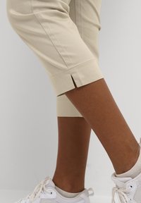 Beige cropped trousers feature a straight cut with a side slit at the hem. Paired with white sneakers, displaying a textured ankle.