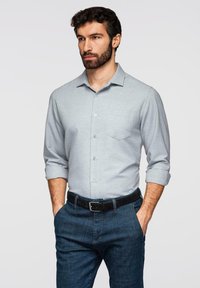 Homme aux cheveux bouclés foncés et à la barbe, portant une chemise grise claire à boutons avec les manches retroussées, un jean foncé et une ceinture noire, les mains dans les poches.