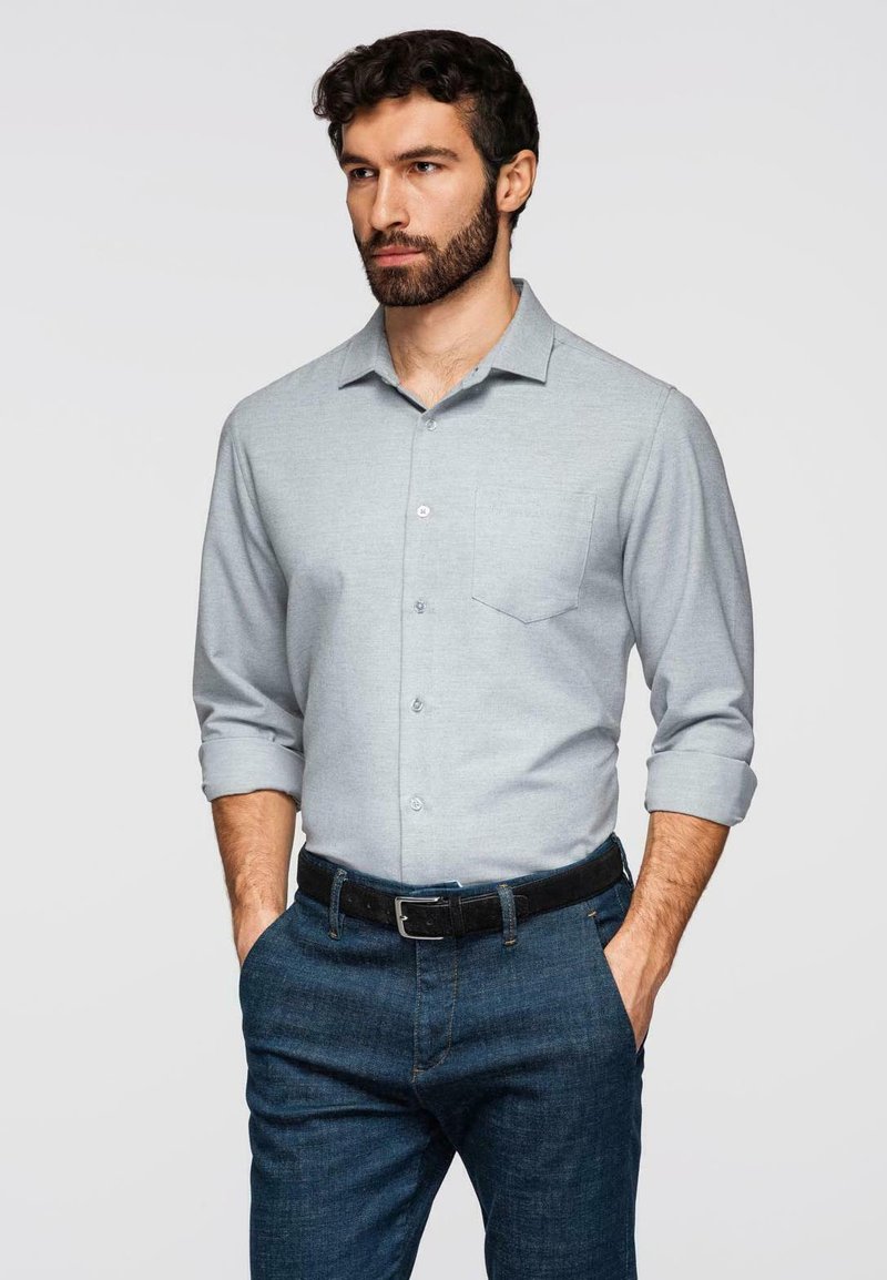 Homme aux cheveux bouclés foncés et à la barbe, portant une chemise grise claire à boutons avec les manches retroussées, un jean foncé et une ceinture noire, les mains dans les poches.