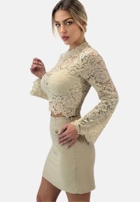 Top in pizzo color beige chiaro con maniche a campana e orlo scallop, abbinato a una mini gonna beige aderente con dettagli di bottoni frontali.