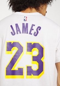 Camiseta de algodón blanca con "JAMES" en texto púrpura encima de "23" en amarillo con contorno púrpura. El logo de la NBA está encima del texto.