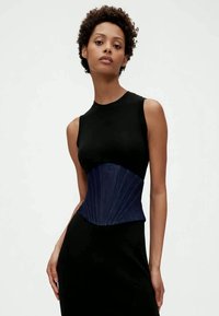 Vrouw met kort krullend haar, gekleed in een mouwloze zwarte jurk en een blauwe korsetachtige riem, staande tegen een effen witte achtergrond.
