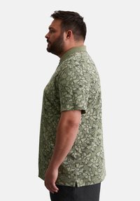 Kurzarm-Polohemd aus olivgrünem Stoff mit floralem Muster, ausgestattet mit einem Kragen und seitlichen Schlitzen. Die strukturierte Oberfläche verleiht dem Shirt eine besondere Note.