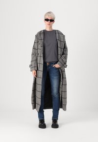 Casaco longo xadrez cinza com grandes lapelas, usado sobre uma blusa cinza escura e jeans azuis. Modelo está de pé com as mãos nos bolsos, usando óculos de sol pretos.