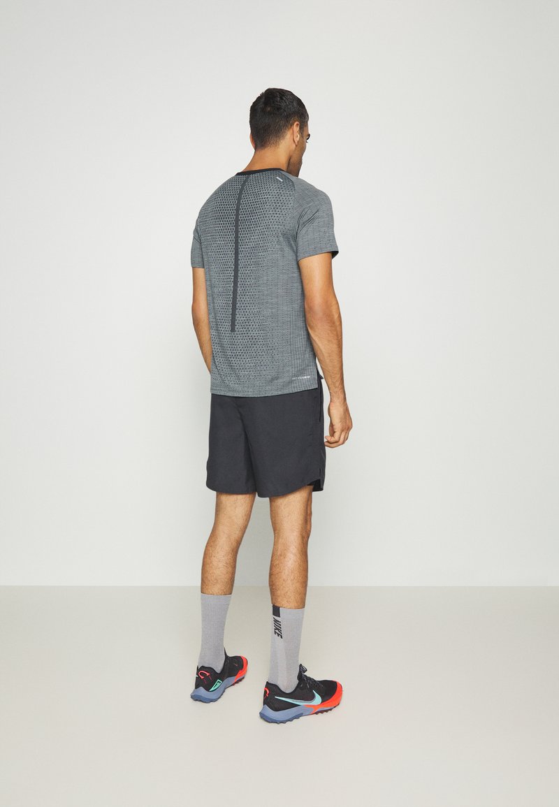 GrauesSportt-Shirt mit einem Mesh-Muster und einer vertikalen Naht auf dem Rücken, kombiniert mit schwarzen Shorts und schwarzen, roten sowie blauen Sneakers.