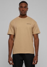 Beige bomulds t-shirt med korte ærmer og rund halsudskæring, som har sort tekst "glemte ansigtter" på venstre brystområde.
