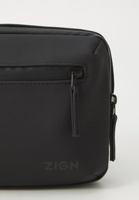 Sac bandoulière noir en matériau synthétique mat, doté d'une poche zippée et d'un logo "ZIGN" en relief à l'avant, au design minimaliste.