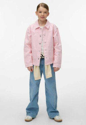 Jauna persona stāv, valkājot rozā jakas ar pogām, baltu kreklu, plašas zilas džinsas, apģērbtu bēša krāsas džemperi apkārt viduklim un baltus tenisa apavus.