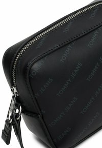 Bolso de cuero sintético negro con cremallera plateada, patrón de logo "TOMMY JEANS" en relieve y forma rectangular con textura suave.