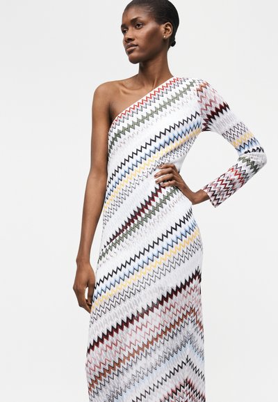 Γυναίκα που φοράει ένα μακρύ φόρεμα με έναν ώμο και σχεδίαση ζιγκ-ζαγκ σε λευκές, μαύρες, κόκκινες, μπλε, κίτρινες και πράσινες αποχρώσεις, πόζα με το χέρι στη μέση.