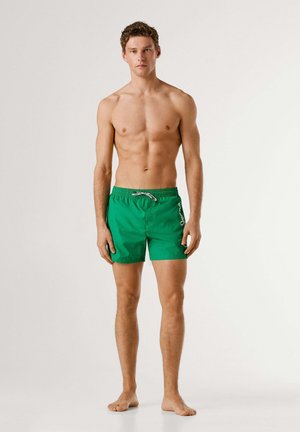 Giovane uomo in piedi a piedi nudi che indossa pantaloncini verdi da bagno con coulisse bianca su sfondo semplice.