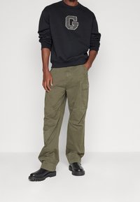 Personne portant un sweat-shirt noir avec un grand logo "G", un pantalon cargo vert olive et des bottes noires épaisses, posant devant un fond blanc.