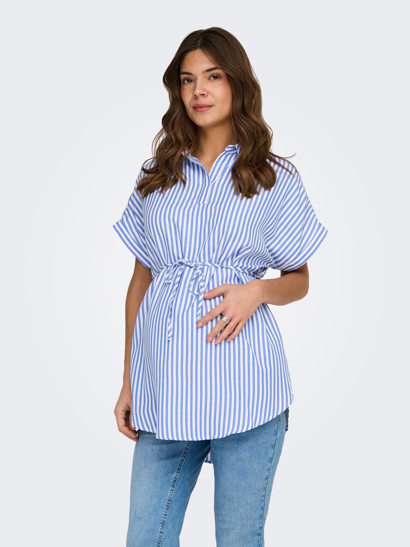 Femme aux cheveux bruns ondulés portant un chemisier de maternité rayé verticalement bleu et blanc avec une ceinture à la taille, assorti à un jean bleu clair.