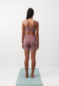 Pehmustettu urheiluliivit ja shortsit hillityssä laventelin sävyssä, joissa on ristikkäiset selkäremmit ja korkea vyötärö, päällä vaaleansinisellä joogamatolla.