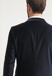 Blazer in velluto blu navy con uno spacco singolo sul retro e rever a punta, che mostra una superficie liscia e morbida.