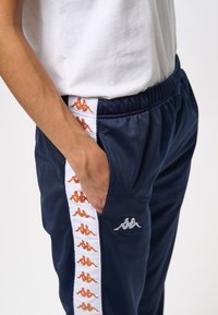 Person klädd i marinblå träningsbyxor med vita och orange logostripes, med handen i fickan, tillsammans med en vit t-shirt.