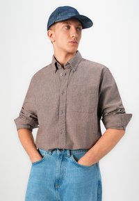 Joven con una camisa marrón de botones y mangas arremangadas, jeans azules y una gorra de cuadros azules, con las manos en los bolsillos, mirando hacia un lado.