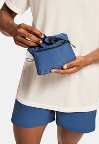 Des mains dépliant une veste compacte pliante bleue de sa pochette zippée, portée sur une chemise blanche et un short bleu.