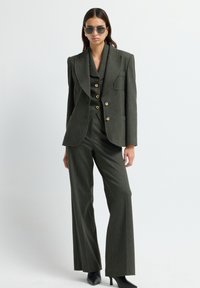 Femme portant un costume sur mesure vert foncé avec des boutons dorés, des chaussures à talons noirs à bout pointu et des lunettes de soleil, se tenant devant un fond blanc uni.