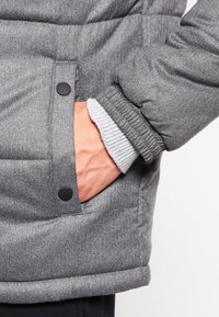 Gråt dunjakke stof med en glat tekstur, der har en frontlomme med sort trykknap og ribbet grå manchet ved ærmet.