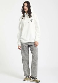 Biała bluza z kapturem ze ściągaczami i logo z przodu, zestawiona z szarymi wzorzystymi spodniami oraz wielokolorowymi sneakersami. Casualowy, luźny krój.