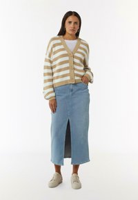 Beige-weißer, gestreifter Cardigan mit V-Ausschnitt und Knopfverschluss. Kombiniert mit einem hellblauen Denim-Rock mit Schlitzen vorne. Neutrale Sneakers.