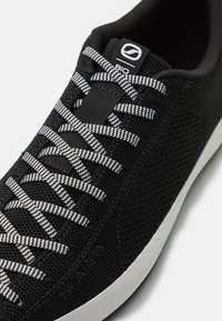 Svart sneakers med en teksturert strikket overdel, med hvite og svarte stripete snøringer, et minimalistisk design, og en hvit såle.