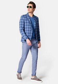 Niebieska marynarka w kratę z wyraźnymi klapami, noszona na niebieskim swetrze z dekoltem w serek, w połączeniu z jasnoniebieskimi spodniami slim fit i szarymi sneakersami. Zielone okulary przeciwsłoneczne.