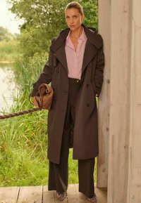 Rjava vodoodporna trench jakna z velikim ovratnikom, gumbom za zapiranje in stranskimi žepi, oblečena čez svetlo roza srajco in temne hlače.