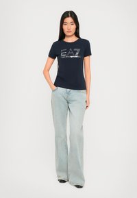 Kvinne med langt mørkt hår som har på seg en marineblå EA7 Emporio Armani t-skjorte og lyseblå vide jeans, står med den ene hånden i lomma.