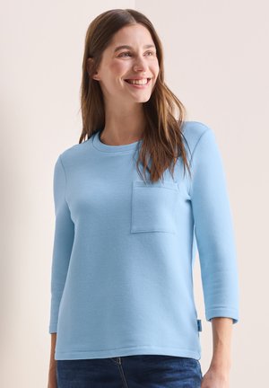 Maglietta azzurra chiara a maniche lunghe con tessuto testurizzato, scollatura rotonda e una piccola tasca sul petto. Il design presenta una vestibilità rilassata.