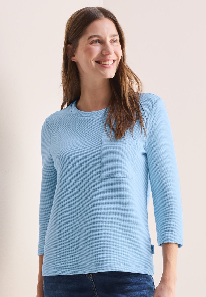 Lichtblauw longsleeve shirt van een textuurstof, met een ronde halslijn en een klein borstzakje. Het ontwerp heeft een relaxte pasvorm.