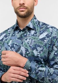 Marineblauwe Shirt met paisleypatroon in groene en blauwe tinten; heeft een puntige kraag, lange mouwen en een knopenlijst. Zachte stoftextuur.