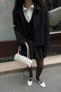 Schwarze Wolljacke über einem schwarzen Top und Rock, kombiniert mit weißen Ballerinas und einer kleinen weißen Handtasche. Gestreifter Kragen ist unter der Jacke sichtbar.