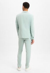 Hellgrüner Zweiteiler aus strukturiertem Stoff, mit schmaler Passform, mittigem Schlitz und passenden Hosen, getragen mit weißen Sneakers.