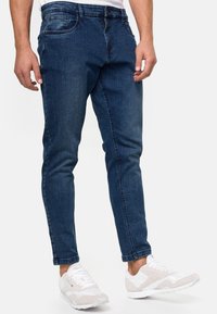 Tummansiniset farkut, joissa on slim fit -malli. Edessä taskut ja vaaleita kulutusoireita. Yhdistetty valkoisiin ja beigeihin urheilukenkiin.