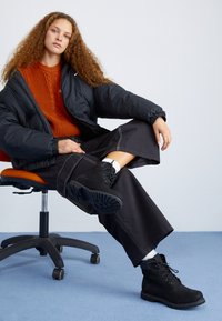 Bottes noires avec une surface texturée, un design à lacets et une semelle robuste. Pull en laine orange chaud et veste noire rembourrée, assis sur une chaise de bureau.