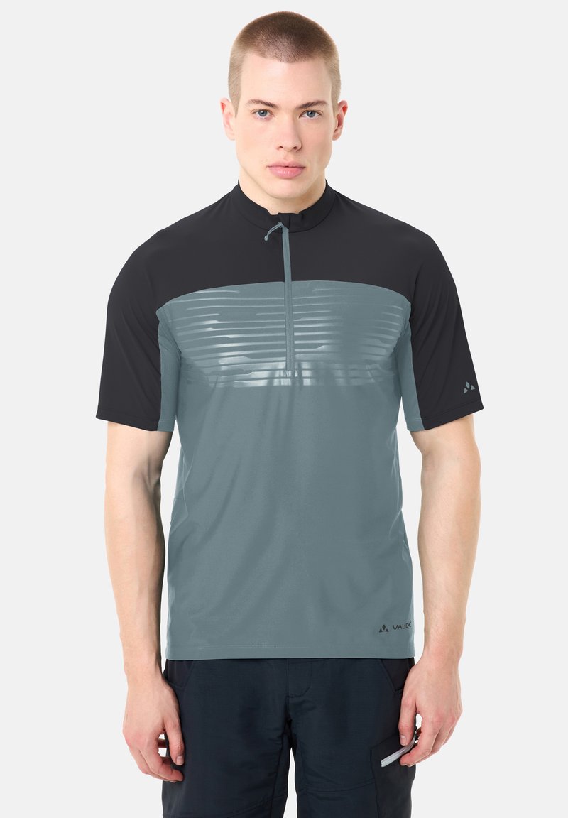 Jeune homme portant un maillot de sport à manches courtes zippé bicolore gris et noir, ainsi qu'un short noir, debout devant un fond uni.