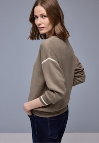 Femme aux cheveux bruns mi-longs portant un pull taupe avec des rayures blanches sur les manches et un jean bleu foncé, regardant par-dessus son épaule.