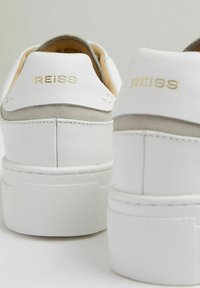Biele kožené tenisky s hladkým povrchom, šedé semišové detaily, textúrovaná gumová podrážka a zlaté logo "REISS" na päte.
