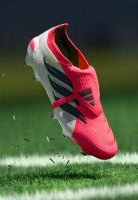 Φωτεινό ροζ και λευκό παπούτσι ποδοσφαίρου με μαύρες ρίγες που σηκώνει γρασίδι σε ένα σημαδεμένο αθλητικό γήπεδο.