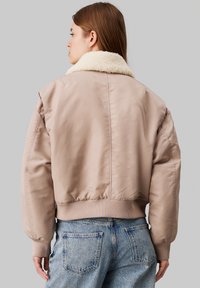 Veste bomber rembourrée en beige clair avec un col en fausse fourrure, des poignets et une ceinture côtelés. Matériau lisse avec une coupe décontractée.